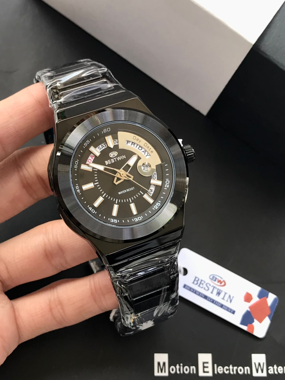 Black wristwatch held by a hand with a visible brand tag on a dark surface
