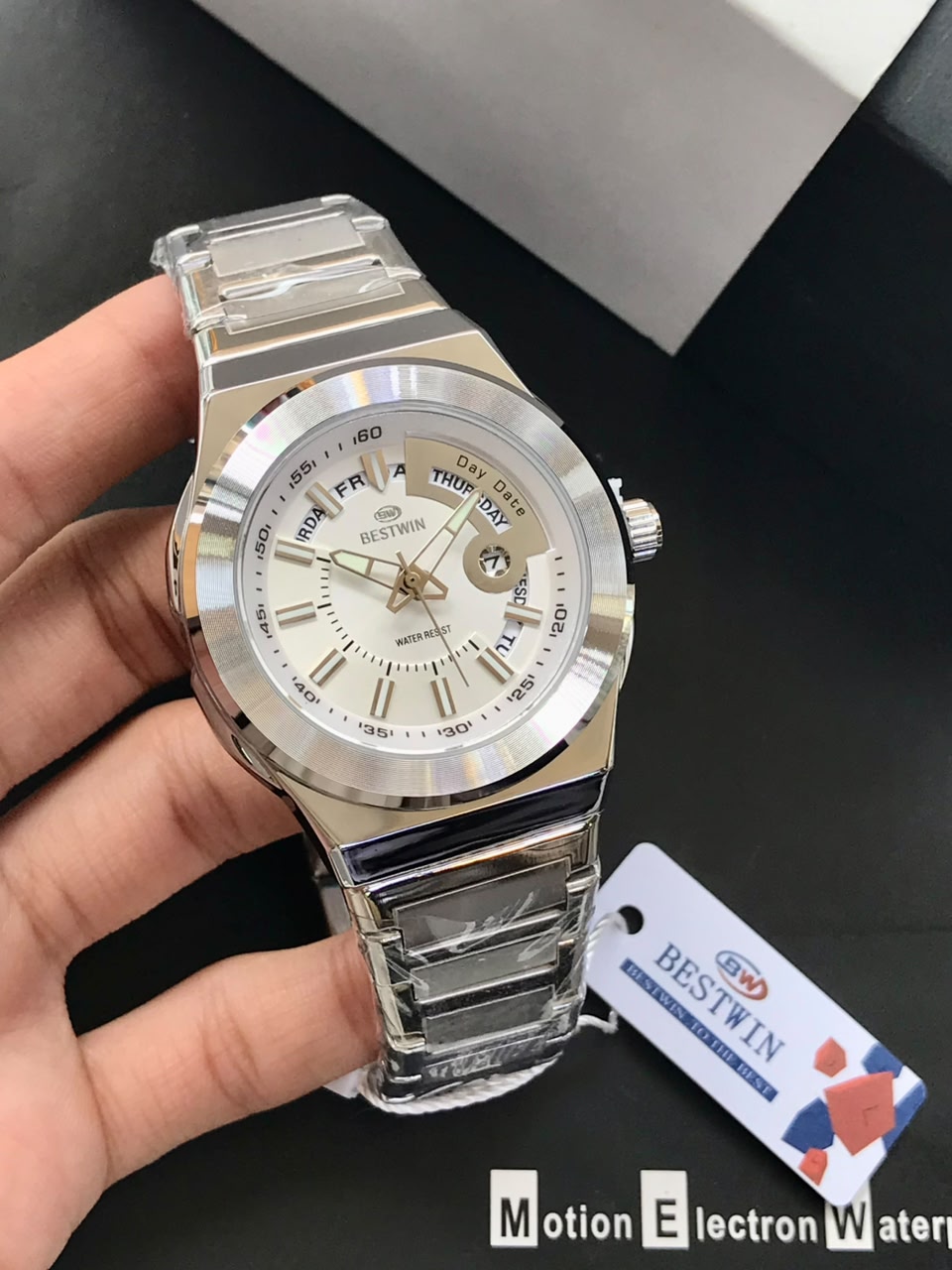 Silver wristwatch held by a hand with a visible brand tag on a dark surface