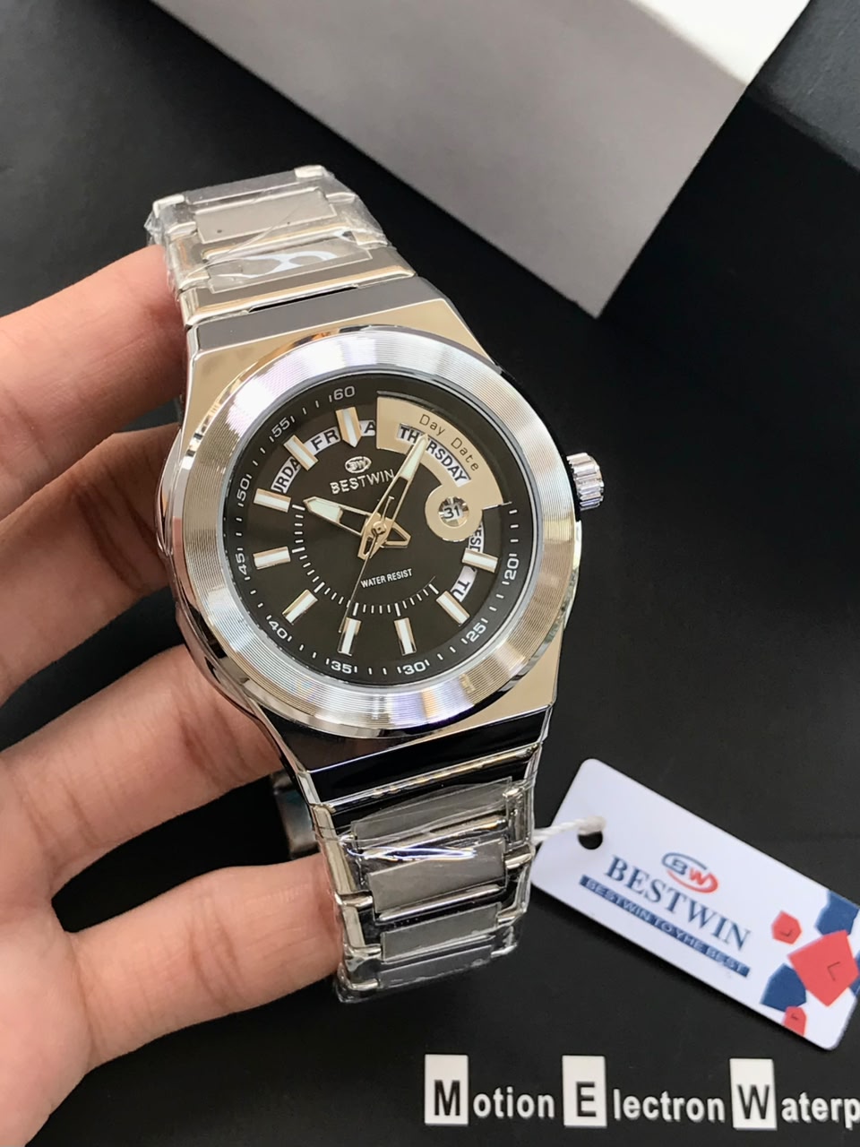 Silver watch with black face held by a hand, brand tag visible
